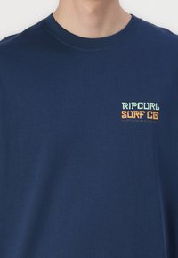 Mørkeblå bomulds T-shirt med rund hals. Udstyret med et flerfarvet "Ripcurl Surf Co" logo på venstre bryst. Glat tekstur, afslappet pasform.