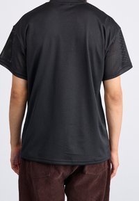 Persona vestida con una camiseta de malla negra holgada de manga corta y pantalones de pana marrones, vista desde atrás sobre un fondo claro liso.