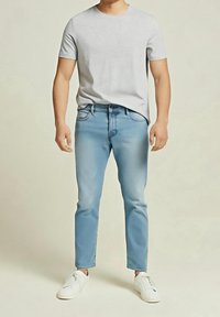 Homme portant une chemise à manches courtes gris clair, un jean bleu clair et des baskets blanches, debout devant un fond neutre.