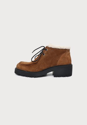 Bottine en daim marron avec lacets noirs, semelle noire épaisse, talon bloc et doublure en polaire blanche visible en haut.