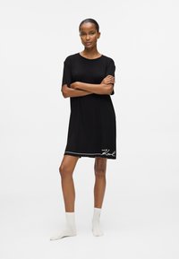 Rochie neagră din bumbac cu mâneci scurte, lungime până la genunchi, cu o margine albă la tiv și numele „Karl” scris cu litere cursive în partea de jos.