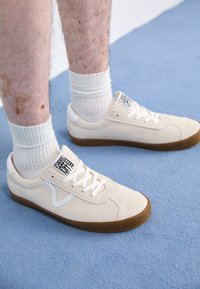 Beige suede sneakers with white accents, a gum rubber sole, and a label on the tongue. Rounded toe and flat laces.