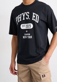 černé bavlněné tričko s bílým nápisem "PHYS. ED" a "EST. 1919", logem "Champion New York" a krátkými rukávy.