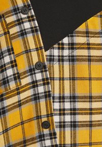 Tissu de chemise à carreaux jaune, noir, blanc et gris avec trois boutons noirs et un vêtement noir en dessous.