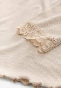 Vaalea beige ribattu kangas, jossa on aaltoileva pitsireuna hihassa, pehmeä tekstuuri hienovaraisilla pystysuorilla viivoilla ja hienovaraisia yksityiskohtia.