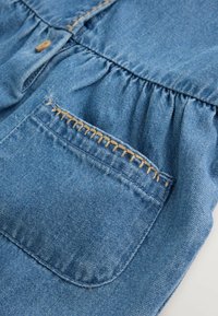 Vêtement en denim bleu avec une poche avant présentant des coutures décoratives beige et un tissu froncé au-dessus de la poche.