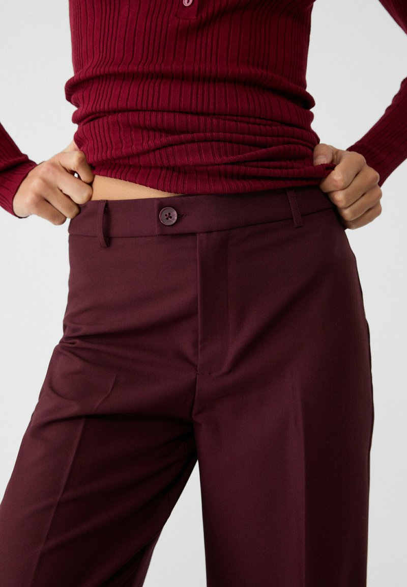 Pantaloni tailored bordeaux con una texture liscia, caratterizzati da una chiusura a bottone singolo, passanti per cintura e tasche frontali per praticità.