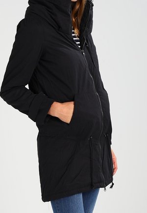 Femme portant un manteau d'hiver noir à capuche avec fermeture éclair et les mains dans les poches, associé à un jean bleu et une chemise rayée en dessous.