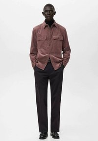 Rostfärgad korduroy-skjorta med två bröstfickor, kombinerad med svarta skräddarsydda byxor och svarta skor, mot en enfärgad vit bakgrund.