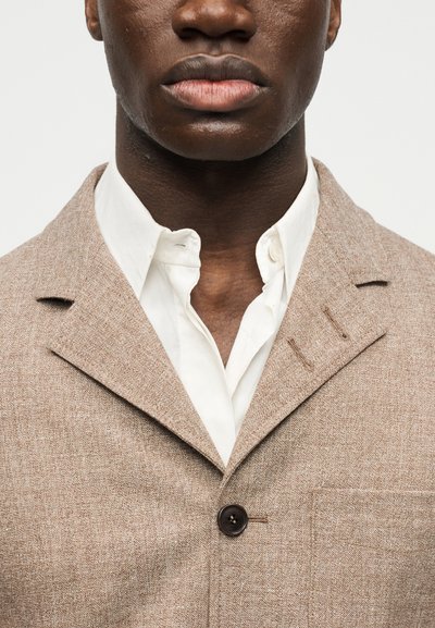 Κοντινό πλάνο προσώπου ατόμου που φοράει μπεζ υφασμάτινο σακάκι πάνω από λευκό πουκάμισο με κουμπιά, εμφανίζοντας τα χείλη, το πηγούνι και το άνω μέρος του στήθους.