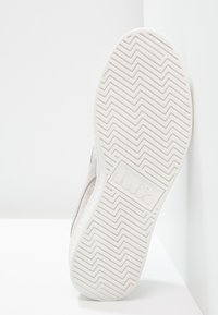 Weißer Sneaker-Sohle mit einem strukturierten Zickzackmuster, das ein dezentes Markenlogo zeigt. Obermaterial scheint aus hell gefärbtem Wildleder zu sein.