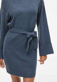 Abito maglione di colore blu scuro, con vita cintrata, maniche lunghe con risvolti ampi e un tessuto strutturato.