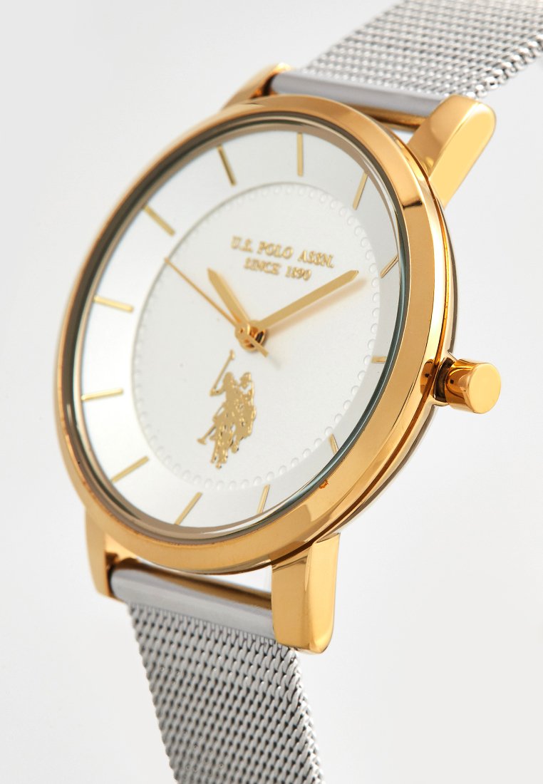 Gouden horloge met een witte wijzerplaat, metalen zilveren band, dunne uurmarkeringen en een logo-embleem. Eenvoudig ontwerp met een ronde kast en helder glas.