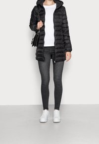 Černá prošívaná bunda s kapucí, se zipem vpředu a dlouhými rukávy, v kombinaci s tmavými džíny a bílými teniskami, doplněná malou kabelkou.