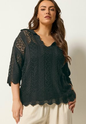 Femme portant un haut noir en crochet ample avec des manches mi-longues jusqu'au coude et un ourlet festonné, assorti à un pantalon de couleur claire.