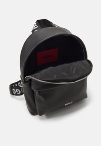 Mochila de cuero negro con forma redondeada, que presenta un bolsillo frontal con cremallera y un interior con patrón. Logotipo "HUGO" en el exterior y etiqueta interior.