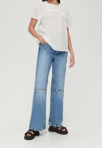 Hvid, struktureret bluse med korte ærmer og sammensnøret halsudskæring, parret med lyseblå brede jeans med slidte knædetaljer og sorte sandaler.