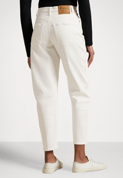 Weiße High-Waist-Jeans mit Gesäßtaschen und Polo-Markenlabel, getragen mit schwarzem Langarmshirt und cremeweißen Sneakers, von hinten gezeigt.