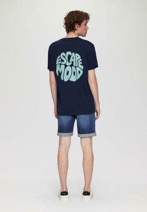 Marineblaue Baumwoll-T-Shirt mit einem hellblauen runden "ESCAPE MOOD"-Grafik auf dem Rücken, kombiniert mit hochgekrempelten Denim-Shorts und schwarzen Slippern.