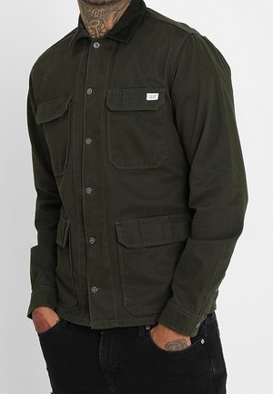 Homme portant une veste boutonnée vert foncé avec poches à rabat sur la poitrine et la taille, mains dans les poches d'un pantalon noir, tatouages visibles au cou et aux mains.
