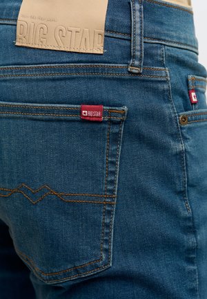 Tmavě modré džíny z denimu s prošívanými detaily. Mají koženou nášivku s nápisem "BIG STAR" v pase a červenou značkovou visačku na zadní kapse.