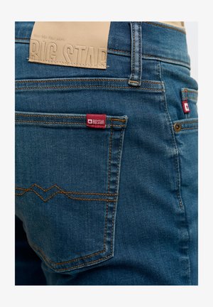 Tmavě modré džíny z denimu s prošívanými detaily. Mají koženou nášivku s nápisem "BIG STAR" v pase a červenou značkovou visačku na zadní kapse.
