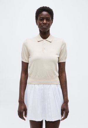 Γυναίκα με κοντά κατσαρά μαλλιά, ντυμένη με μπεζ πόλο μπλούζα με κοντά μανίκια και λευκή πλισέ φούστα, στέκεται μπροστά σε απλό φόντο.