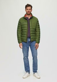 Grüne Pufferjacke mit vertikalen Nähten, Reißverschluss-Tasche und geripptem Kragen; getragen über einem dunklen Hemd und kombiniert mit blauen Jeans und weißen Sneakers.