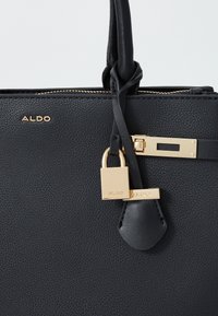 Sac à main en simili cuir noir avec quincaillerie dorée, mettant en avant un détail en forme de cadenas et un logo, de forme structurée et d'une texture lisse.
