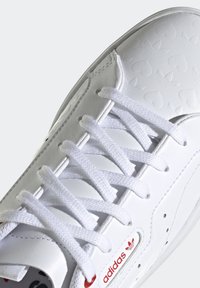 Vita lädersneaker med texturerat hjärtmönster, vita snören och röda logodetaljer. Har en rund tå och vadderad krage.