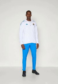 Valkoinen vetoketjullinen takki sinisillä yksityiskohdilla, jossa on logo ja raidalliset hihat, yhdistettynä sinisiin housuihin ja mustiin urheilujalkineisiin.