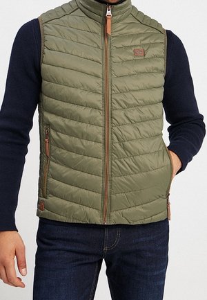 Homme portant une veste matelassée sans manches vert olive avec fermeture éclair à l'avant et une chemise à manches longues bleu marine, les mains dans les poches.