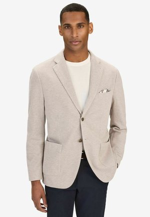 Man i beige texturerad kavaj över vit skjorta med mönstrad näsduk, mörka byxor, står med ena handen i fickan.