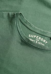 T-shirt en coton vert avec un col rond. L'étiquette indique "SUPERDRY CO. CRAFT & DETAIL" en blanc. Texture douce avec une finition lisse.