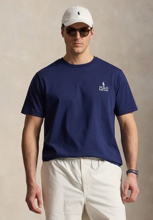 Homme portant un t-shirt Polo Ralph Lauren bleu marine, une casquette blanche, des lunettes de soleil, un pantalon blanc et une montre-bracelet avec le logo Polo, debout devant un fond uni.