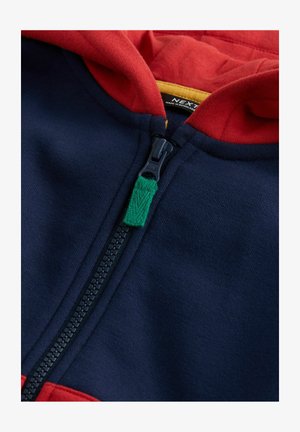 Jachetă cu glugă, cu un corp de culoare bleumarin și glugă roșie, cu un fermoar negru și un tab de mână din țesătură verde. Textură moale și design modern.