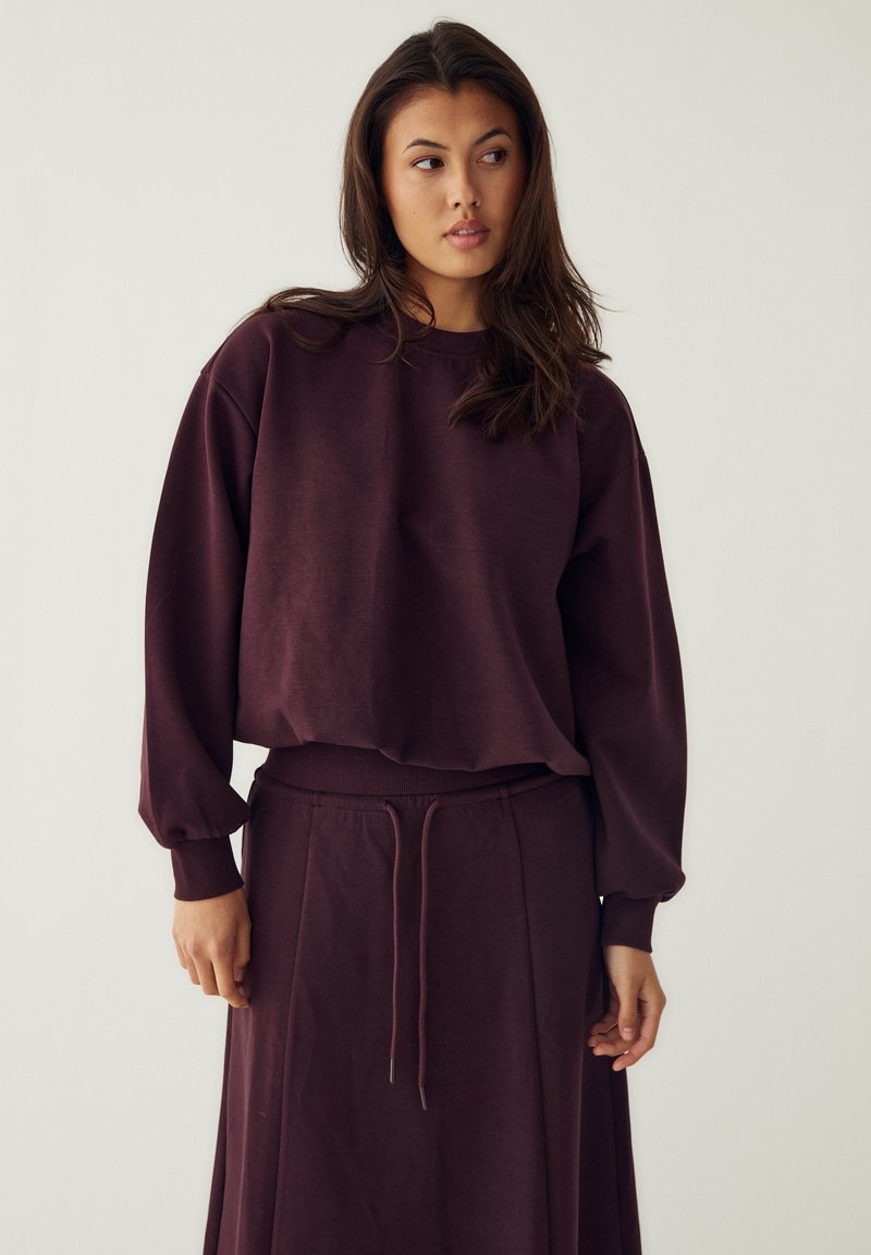 Femme aux longs cheveux foncés portant un haut ample violet foncé à manches longues et une jupe assortie à cordon, sur un fond clair uni.