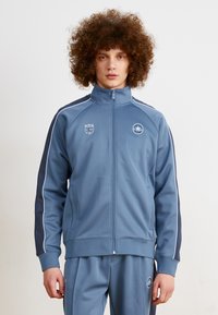 Chaqueta deportiva azul con un cuello alto, franjas marinas en contraste y cierre de cremallera. Presenta logos en el pecho y la manga.