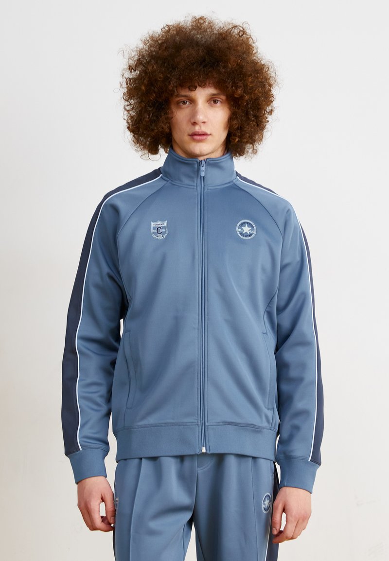 Chaqueta deportiva azul con un cuello alto, franjas marinas en contraste y cierre de cremallera. Presenta logos en el pecho y la manga.