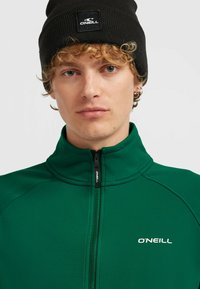 Grüne Zip-Jacke mit hohem Kragen und O'Neill-Logo. Kombiniert mit einer schwarzen gerippten Mütze, die ein quadratisches Logo-Patch zeigt.