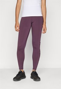 Vrouw in paars leggings met gestreepte zijkanten, witte top en zwarte sportschoenen, staande tegen een effen lichte achtergrond.