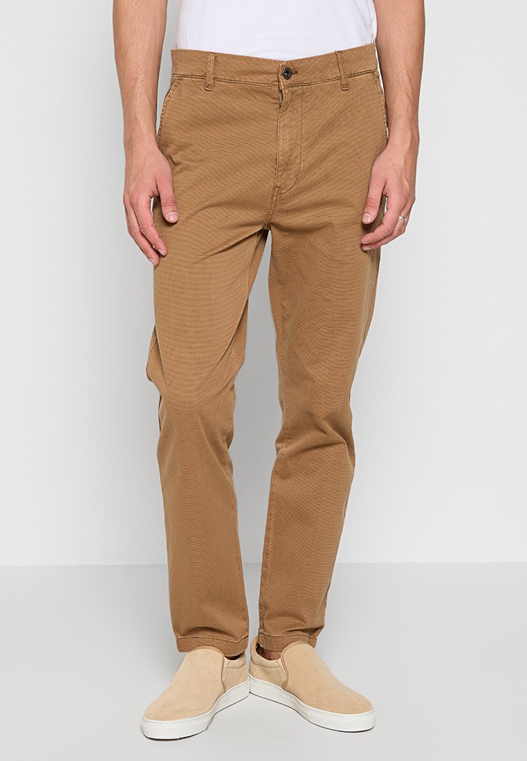 Beige Chinos aus strukturiertem Baumwollstoff, mit Knopfverschluss, Fronttaschen und schmalem Schnitt. Getragen mit hellen Slippern.