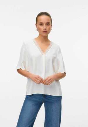 Hvid bluse med V-udskæring og blonderDetaljer langs halsen. Korte, løse ærmer og glat tekstur. Matchet med blå denim jeans.