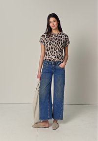 Femme portant un haut à manches courtes imprimé léopard, un jean bleu à jambes larges, des chaussures beige à enfiler, tenant une veste beige dans une main.