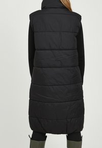 Černá péřová vesta s vysokým límcem, prošívaným designem a prodlouženým tvarem, který se vyznačuje hladkým povrchem a bez viditelného kování.