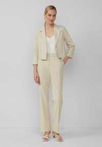 Beige cropped blazer met een enkele knoop, gecombineerd met bijpassende tailleregisterbroek. Gladde textuur, slim fit, gedragen met witte sandalen met bandjes.