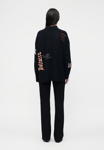 Femme de dos portant un pull noir surdimensionné avec des patchs de texte rouges et blancs, ainsi qu'un pantalon noir, sur un fond uni.