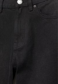 Gros plan d'un jean en denim noir montrant le bouton avant, la zone de la fermeture éclair et la poche droite avec des détails de couture visibles.
