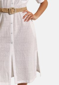 Witte oogjesjurk met knoopsluiting, korte mouwen en een zijsplit. Gemaakt met een beige gevlochten riem met een ronde gesp.