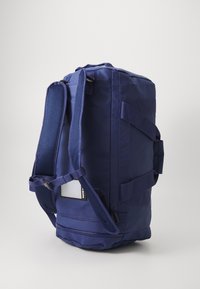 Plavi duffle bag od izdržljive, teksturirane tkanine. Ima podesive podstavljene naramenice i bočnu ručku, s minimalnim hardverom.